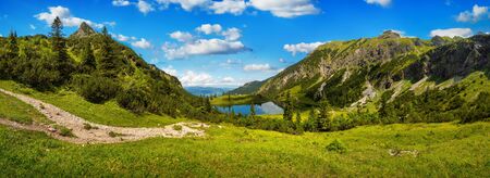 Gorgeous mountain range surrounding a lake, with deep blue sunny sky and green meadows in the foregroundの写真素材
