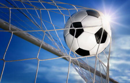 Football or soccer goal, a ball with classic design flying into the net, with blue sky and sun in the backgroundの写真素材