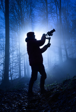 Silhouette of a videographer in a misty forest, dramatically backlit with cool blue color and rays of light behind himの写真素材