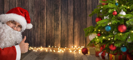 Winking Santa shows thumbs up and presents your text or content on a dark wood background copyspace between himself, a Christmas tree and glowing lightsの写真素材