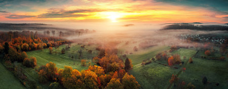 Aerial panorama of a misty rural landscape at sunrise, with colorful autumn trees and green meadows, colorful dramatic sky and layers of mistの写真素材