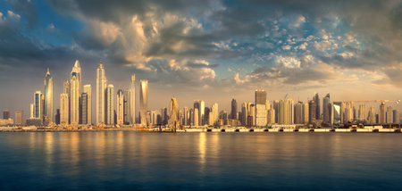 View of the Dubai skyline at sunset from the sea, with the buildings in a row on the horizon reflected in the water. Dramatic clouds and warm sunlit earth colors.の写真素材