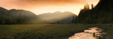 Rays of sunlight falling through mountains after sunset. A panoramic landscape in warm colors with grasslands and a stream in the German Alps.の写真素材