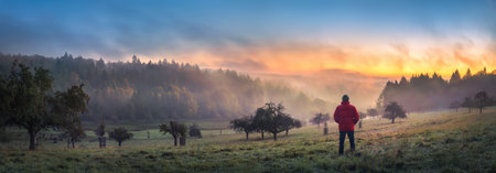 Man standing on a misty hill at sunrise watches the beautiful orange glow in winter. A panoramic...