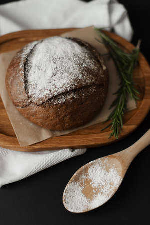 Round black bread on baking paper and a wooden spoon with flour on a black background. Homemade...
