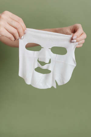 A woman's hands hold a cosmetic face mask made of white fabric. Cosmetic face mask on a green background.の写真素材