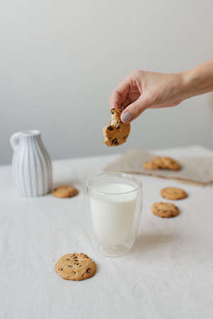 A woman's hand dips a cookie into a glass of milk. Glass glass with milk and cookies on the table.の写真素材