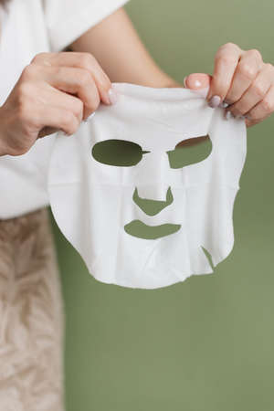 A woman's hands hold a cosmetic face mask made of white fabric. Cosmetic face mask on a green background.の写真素材