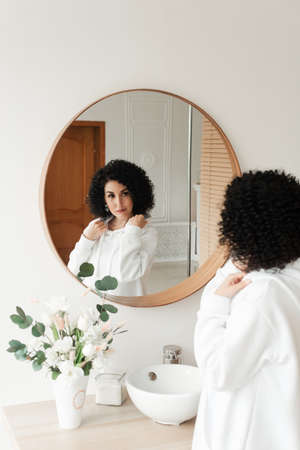 A brunette woman with African curls in a white sweater smiles at herself in the mirrorの写真素材