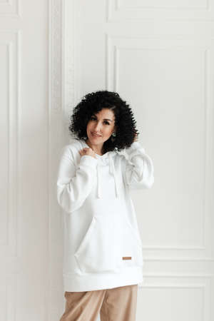 Woman with black curly hair in a white sweater with a hood stands against the background of a white wallの写真素材