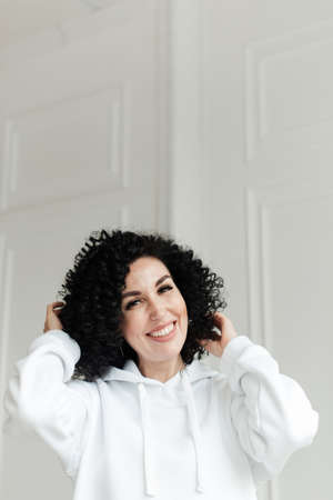 Woman with black curly hair in a white sweater against a white wall laughs and touches her hair