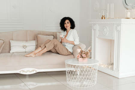 A woman with curly hair sits on the couch barefoot and holds a glass of water against the backdrop...