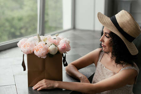 Woman in beige dress and straw hat looks at white and pink bouquet of flowersの写真素材