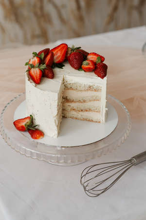 sweet white cream cake with strawberries on a tray and whisk on a white tableの写真素材