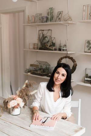 stylish beautiful brunette in white shirt reading a magazine at a wooden table in a stylish roomの写真素材