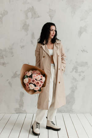 beautiful brunette girl in a beige long trench coat and big boots with a bouquet of flowers in her handsの写真素材