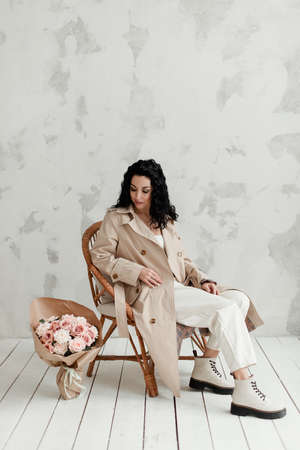 beautiful brunette girl in a beige long trench coat white pants and big boots sits on an armchair with a bouquet of flowersの写真素材
