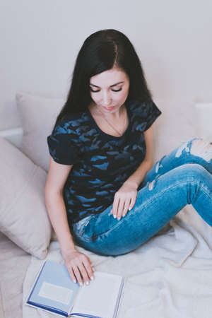 young brunette girl in a t-shirt and blue jeans reads a book on the bedの写真素材