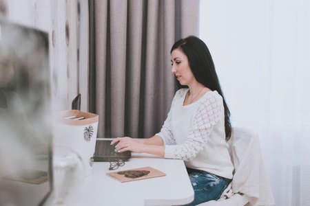 a young brunette girl in a white sweater works at home behind a laptop on a white tableの写真素材