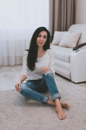 pretty brunette girl in white sweater and blue jeans sitting on the floor at homeの写真素材