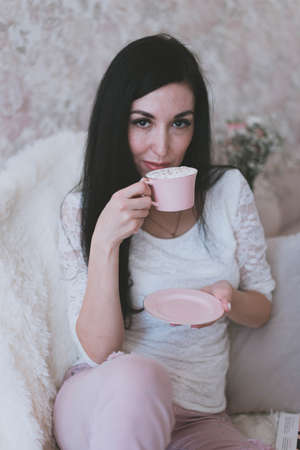 stylish young brunette girl in pink pants and a white sweater holds a mug and saucer in her handsの写真素材