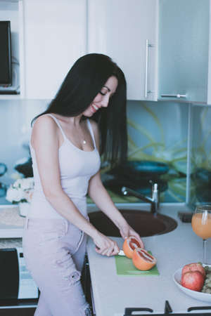 young brunette girl in a purple top cuts a grapefruit with a knife in the kitchenの写真素材