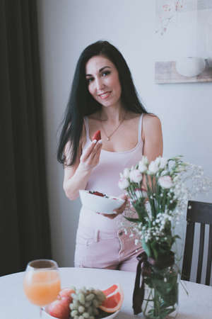 beautiful brunette girl in top eating strawberries and other fruits at the tableの写真素材