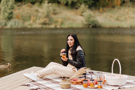 stylish brunette girl in a leather jacket drinks tea from a mug on the lakeside picnicの写真素材
