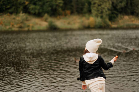 stylish little girl in a hat and black jacket walks near the lake in autumnの写真素材