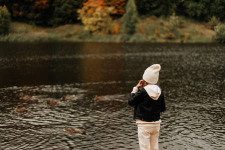 stylish little girl in a hat and black jacket on a background of water and autumn landscapeの写真素材