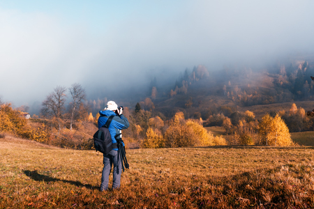 Photographer taking photo of autumn landscape with foggy peaks and orange trees. Ukrainian Carpathians mountainsの写真素材
