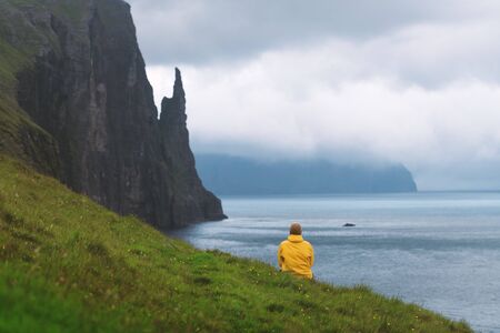 Tourist in yellow jacket looks at Witches Finger cliffs from Trollkonufingur viewpoint. Vagar island, Faroe Islands, Denmark.の写真素材