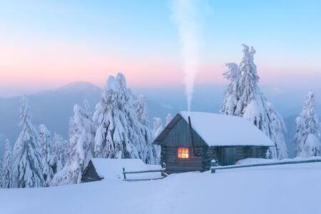 Fantastic winter landscape with wooden house in snowy mountains. Smoke comes from the chimney of snow covered hut. Christmas holiday and winter vacations conceptの写真素材