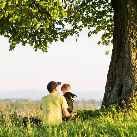 Dad with son in the spring meadow sitting under the tree in tall grass. Travel with child conceptの写真素材