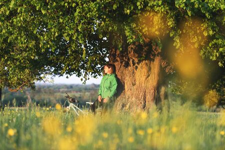 A lonely girl sits under a big old tree near a bicycle. Green summer landscape on city contrsideの写真素材