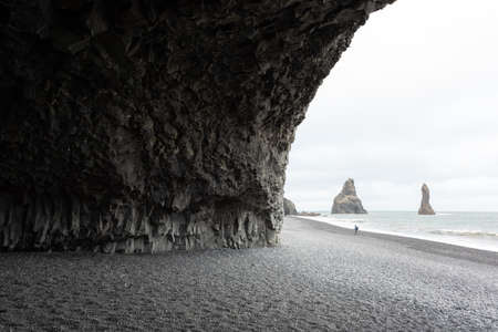 Incredible view on Black beach and Troll toes cliffs in cloudy weather. Lonely tourist with tripod going on coastline. Reynisdrangar, Vik, Iceland. Landscape photographyの写真素材