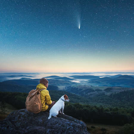 Alone tourist sitting on the edge of the cliff with white dog against the backdrop of an incredible mountains with starry night sky. Landscape photographyの写真素材