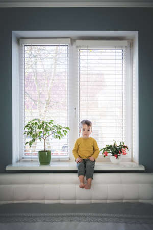 Little loneliness kid boy sitting on windowsill near large window in his room. Childhood conceptの写真素材