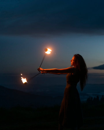 Graceful elegant red haired female fireshow performer making fire dance with two metal fans lit with flames on night mountains meadowの写真素材