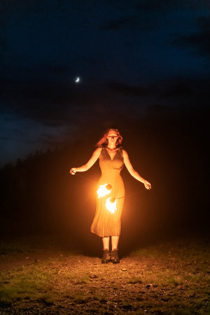 Graceful elegant red haired female fireshow performer making fire dance with two metal fans lit with...