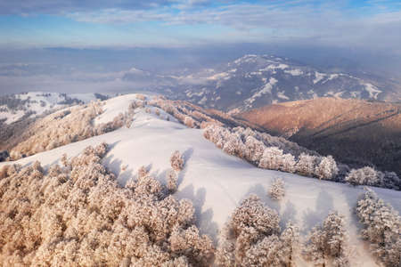Amazing aerial view of mountains range, meadows and snow-capped peaks in winter time. Forest with frost glowing with bright warm sunrise lightの写真素材