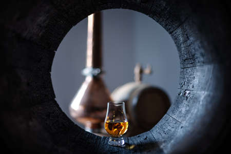 A glass of whiskey in old oak barrel. Copper alambic on background. Traditional alcohol distillery conceptの写真素材