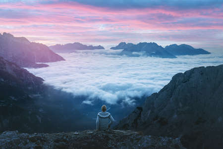 A tourist sitting over the fog at the edge of a cliff in the Dolomites mountainsの写真素材