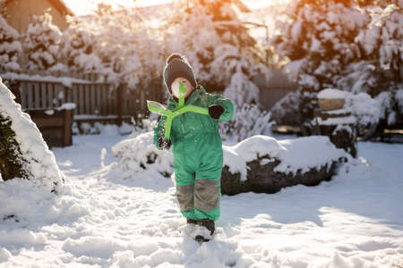 Little child in green jacket and knitted hat make snowballs near houseの写真素材