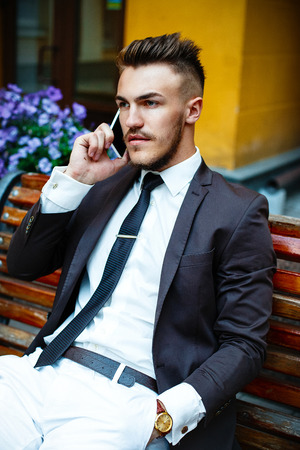 Handsome stylish man in elegant black coat and white shirt holding telephone outdoorsの写真素材