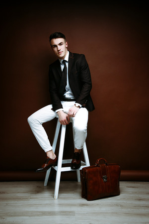 Portrait of handsome stylish man in elegant black suit and white pants sitting on chairの写真素材
