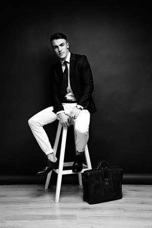 Portrait of handsome stylish man in elegant black suit and white pants sitting on chairの写真素材