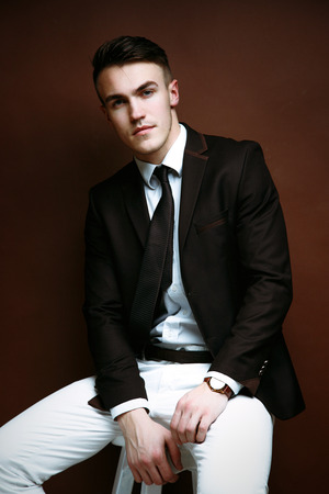 Portrait of handsome stylish man in elegant black suit and white pants sitting on chairの写真素材
