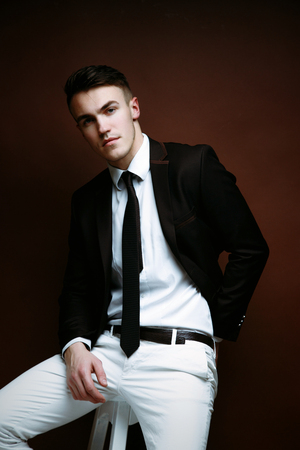 Portrait of handsome stylish man in elegant black suit and white pants sitting on chairの写真素材