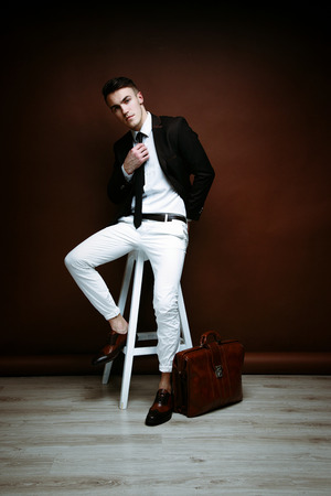 Portrait of handsome stylish man in elegant black suit and white pants sitting on chairの写真素材